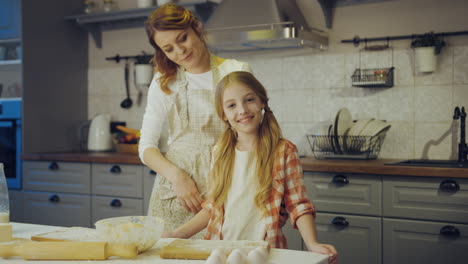 Portrait-shot-of-the-beautiful-mother-hugging-her-pretty-little-daughter-and-they-both-smiling-the-camera-in-the-kitchen-at-the-table-while-baking.-Inside